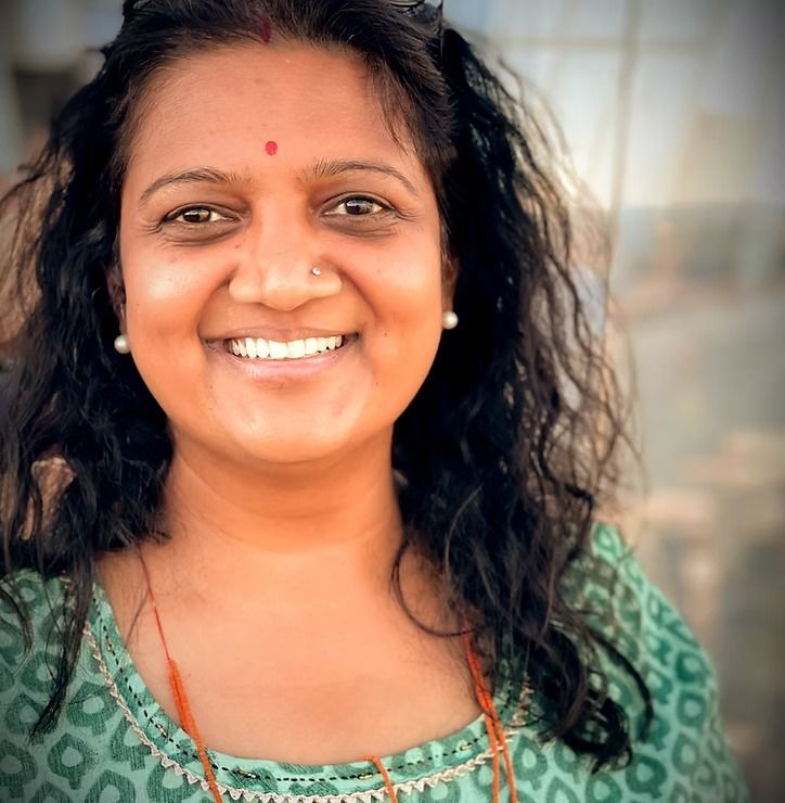 Smiling woman in a green patterned top with curly hair and a red bindi, standing outdoors.