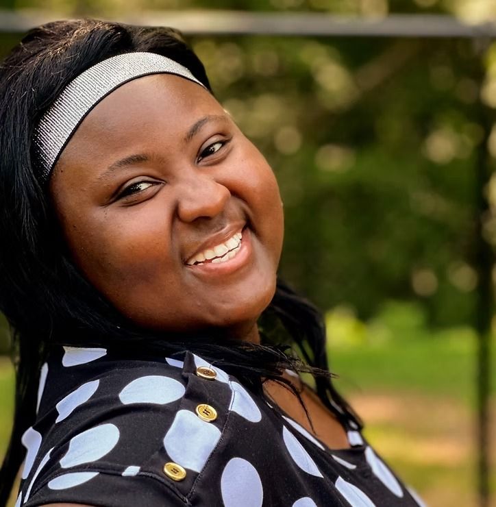 Smiling person in a black-and-white patterned shirt and headband outdoors, with blurred greenery behind them