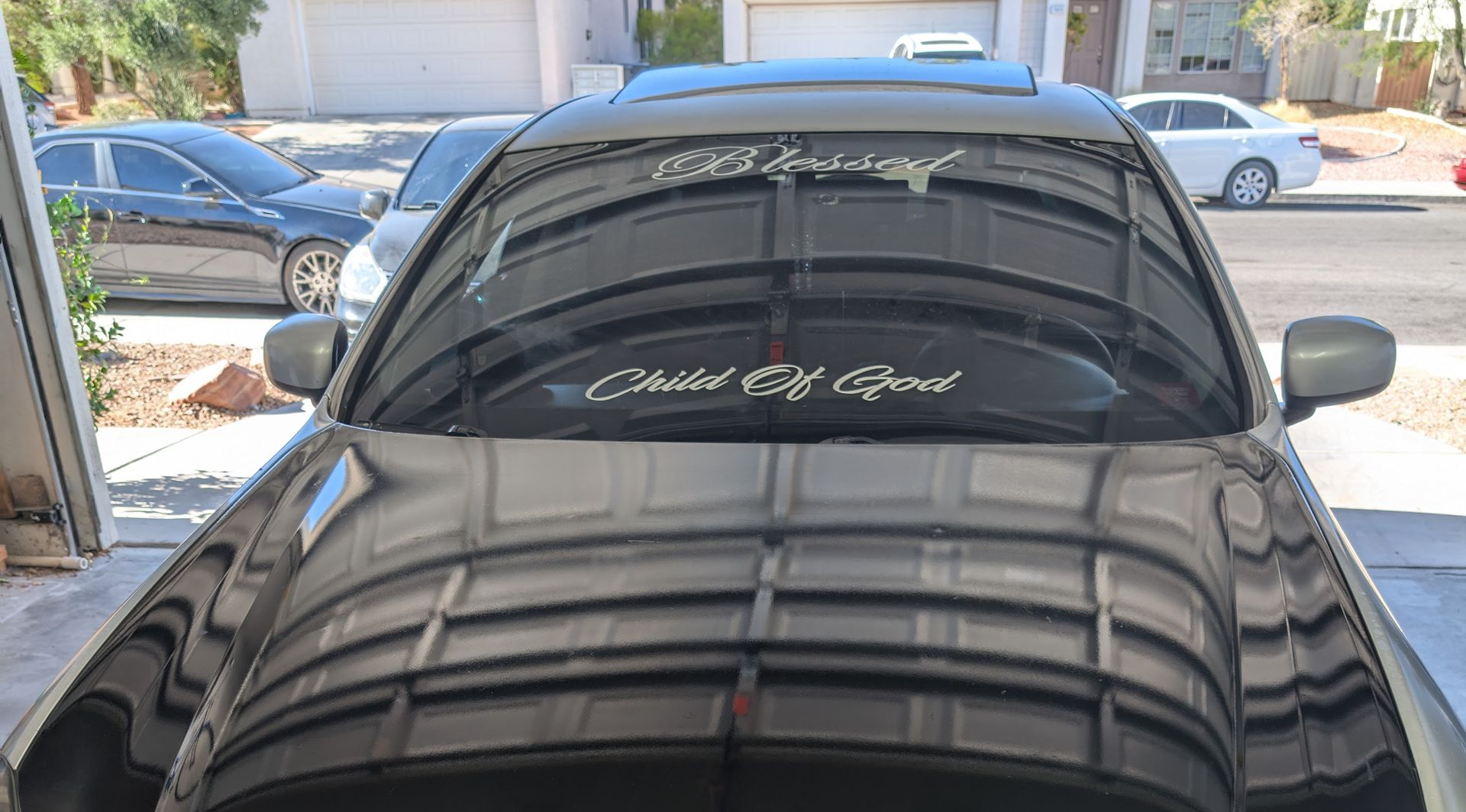 Black car parked in a driveway, front view with reflective hood and windshield sticker