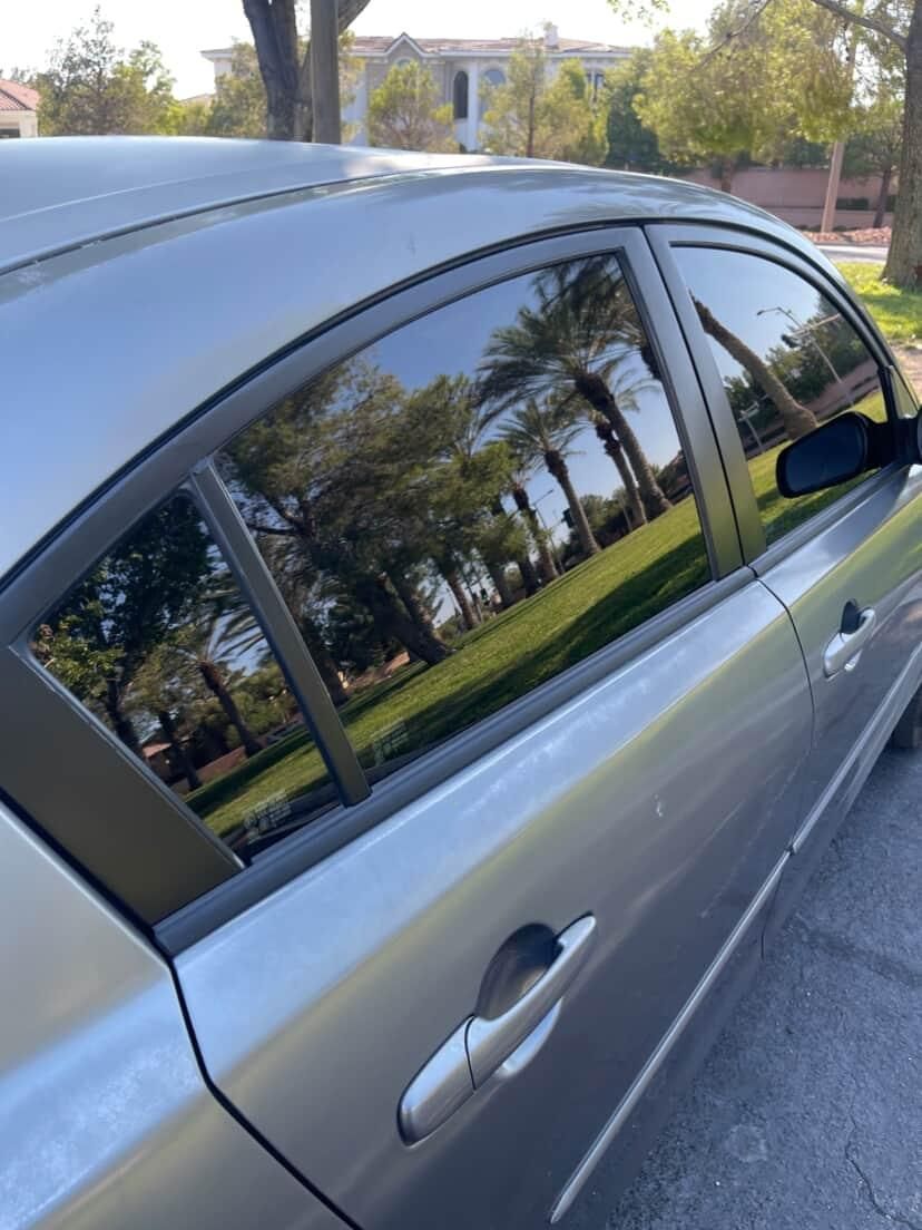 Silver car parked outdoors with tinted windows reflecting palm trees and a grassy area.