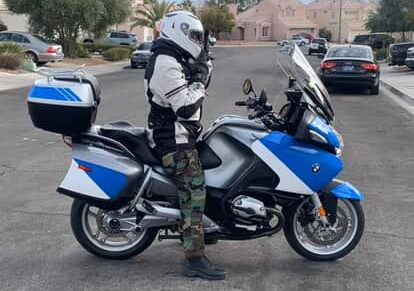Motorcyclist in white helmet and gear standing beside a blue touring motorcycle on a suburban street