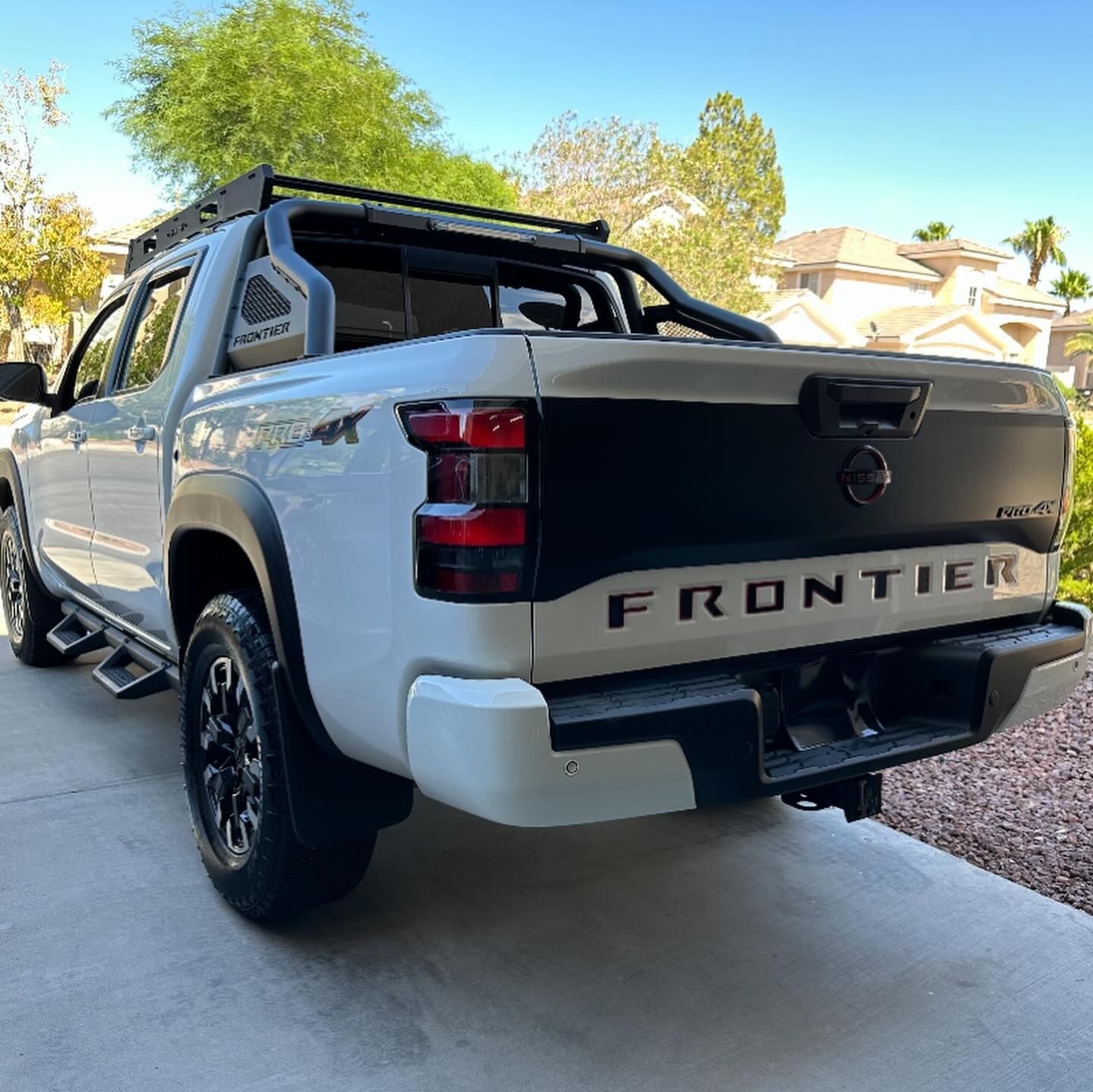 White Nissan Frontier pickup truck parked in a driveway, rear three-quarter view, with black bed rack.