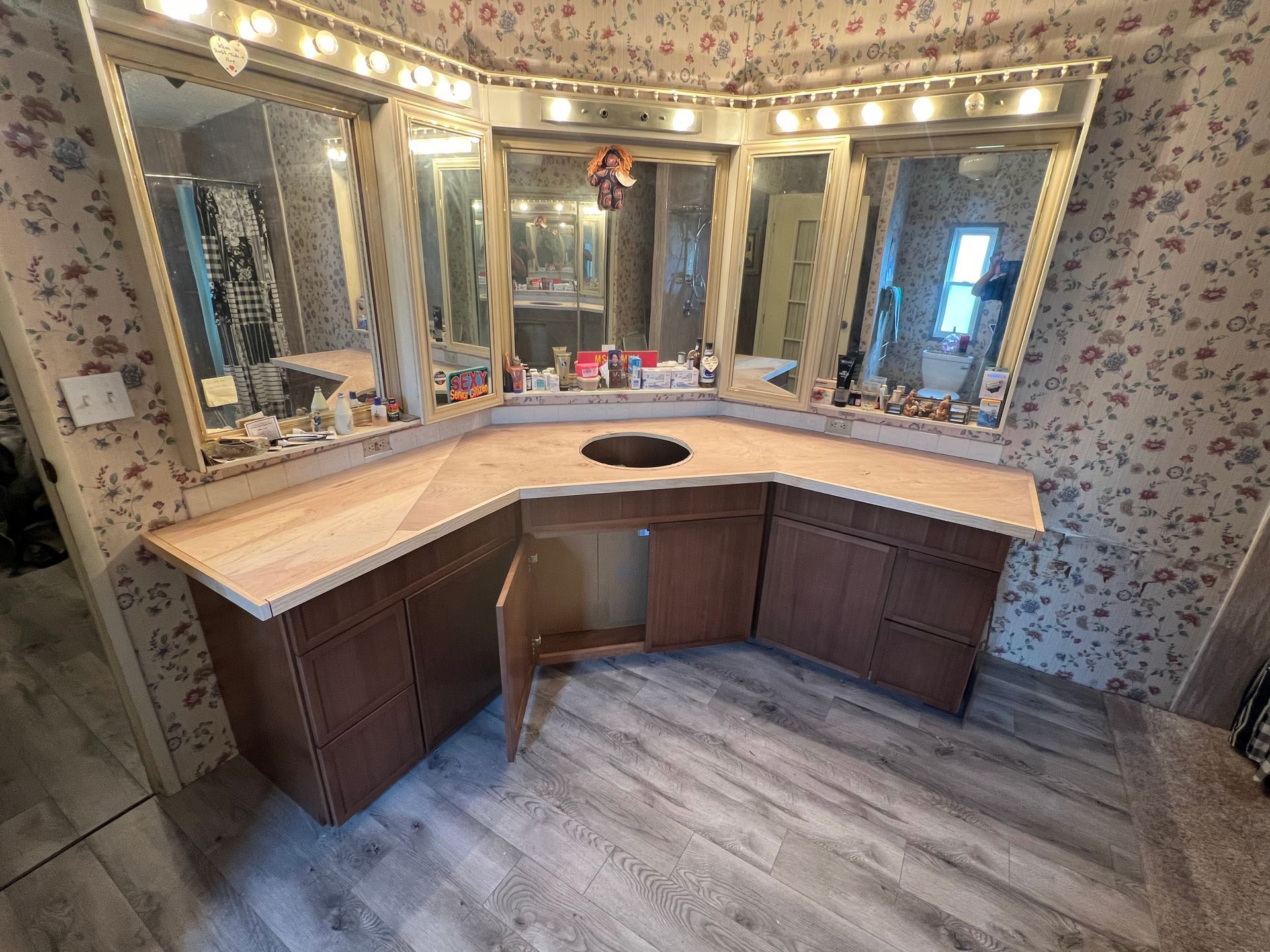 Empty bathroom vanity with lights, mirrors, floral wallpaper, and a curved wood cabinet under a beige countertop