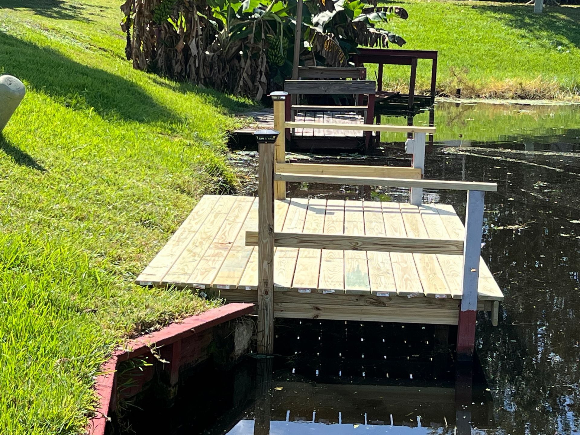 Wooden dock with benches beside a calm pond, bordered by bright green grass and trees