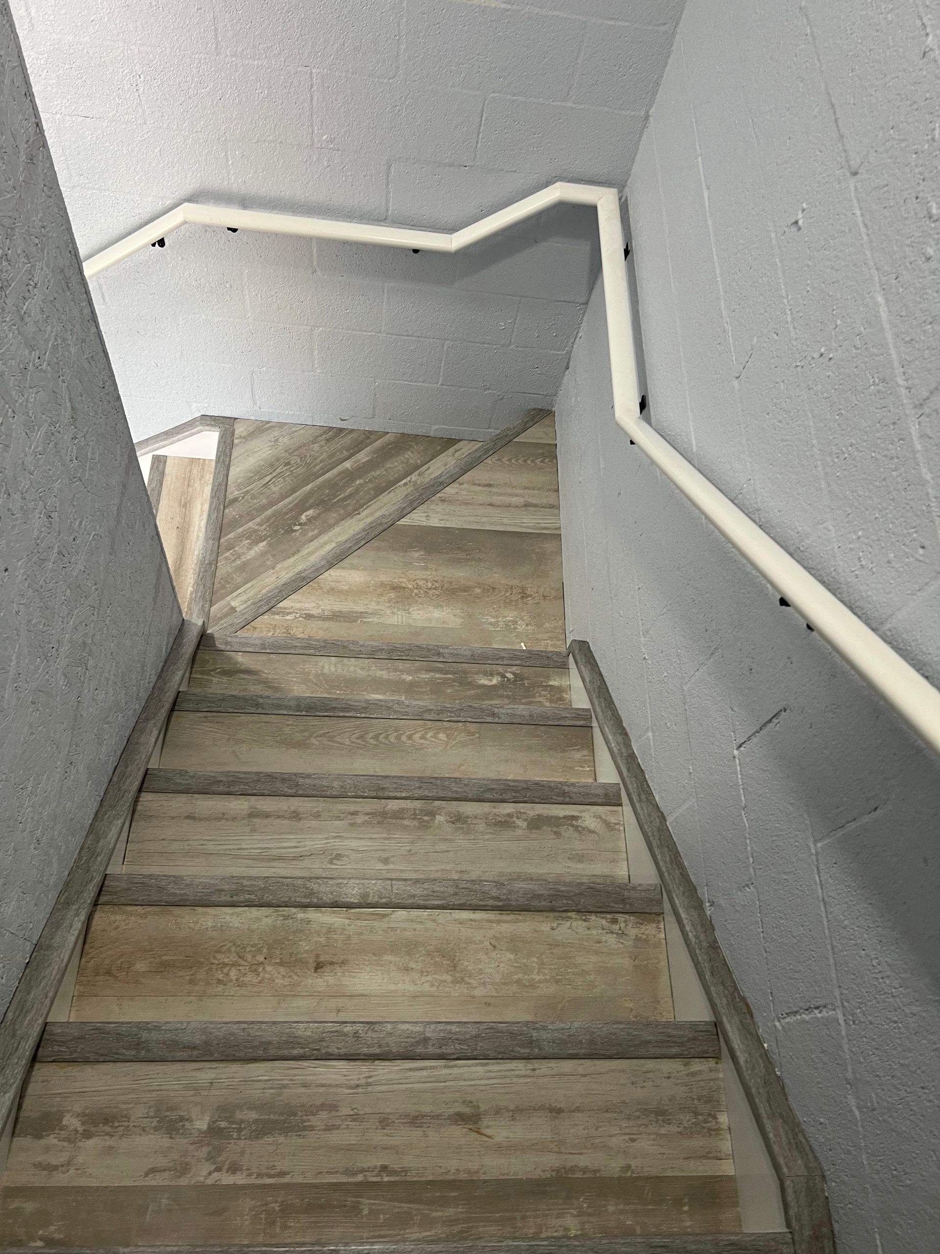 Narrow stairwell with worn wooden steps and gray walls, descending with a white handrail on both sides