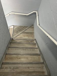 Narrow stairwell with worn wooden steps and gray walls, descending with a white handrail on both sides