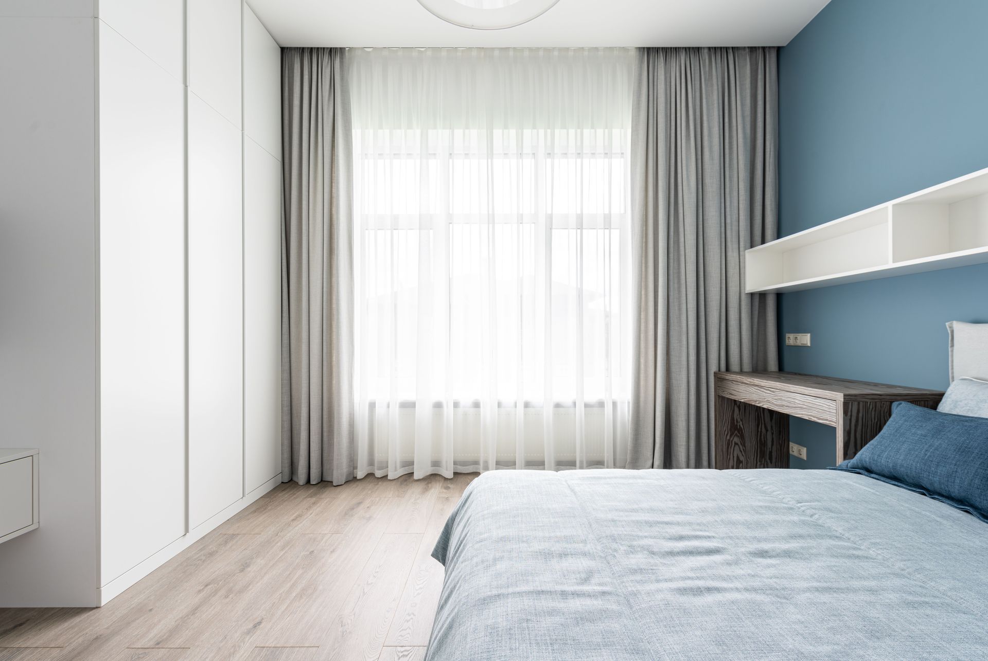Bright modern bedroom with blue accent wall, bed, white wardrobe, and large window with sheer curtains