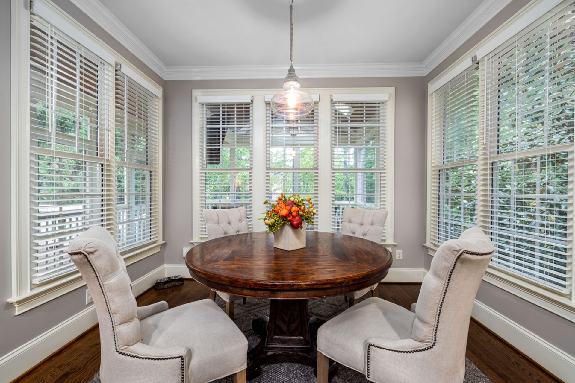 Bright breakfast nook with round wooden table, white chairs, and floral centerpiece beside large windows