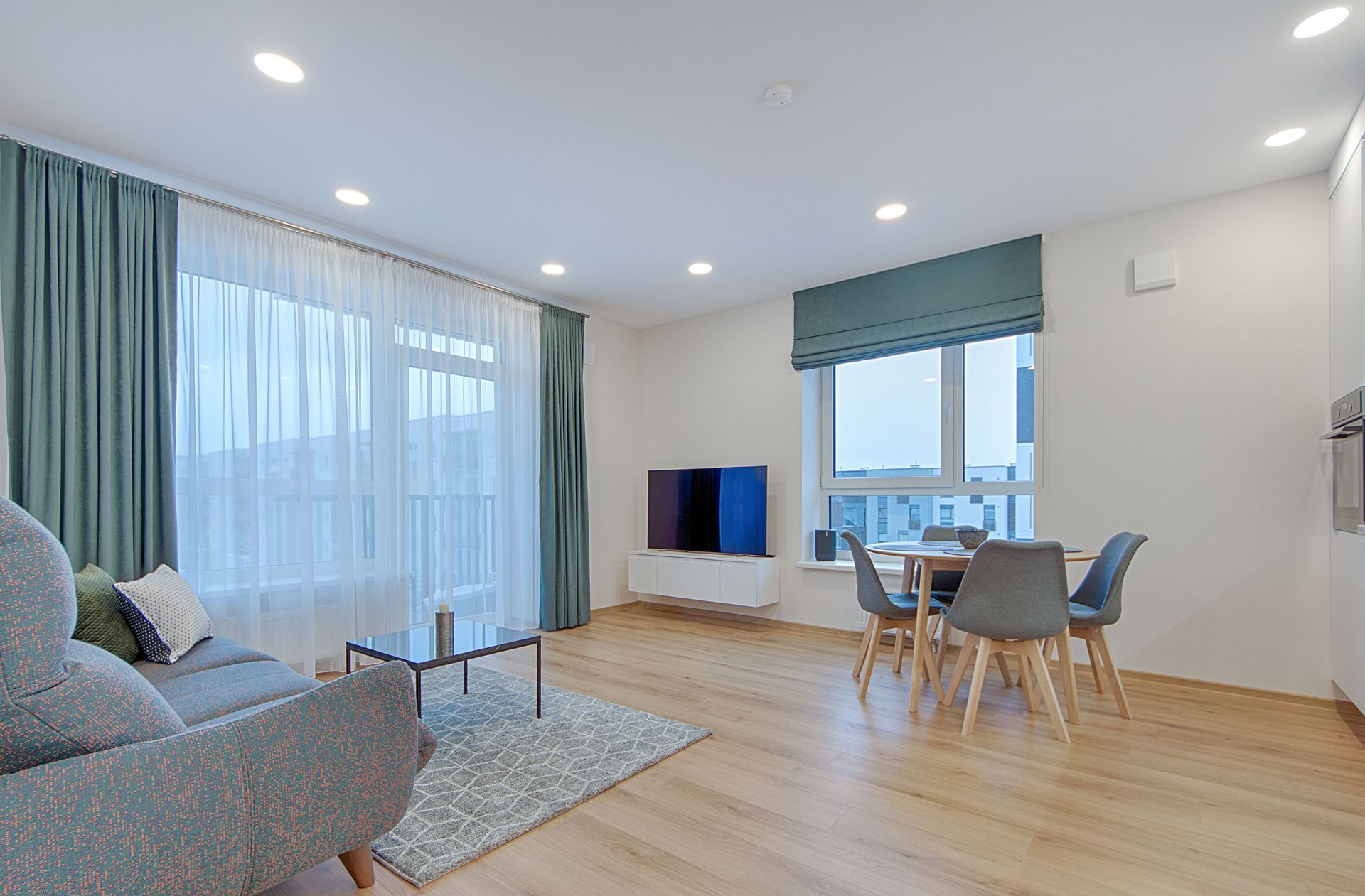 Bright modern living room with gray sofa, TV, dining table, and large windows with blue curtains