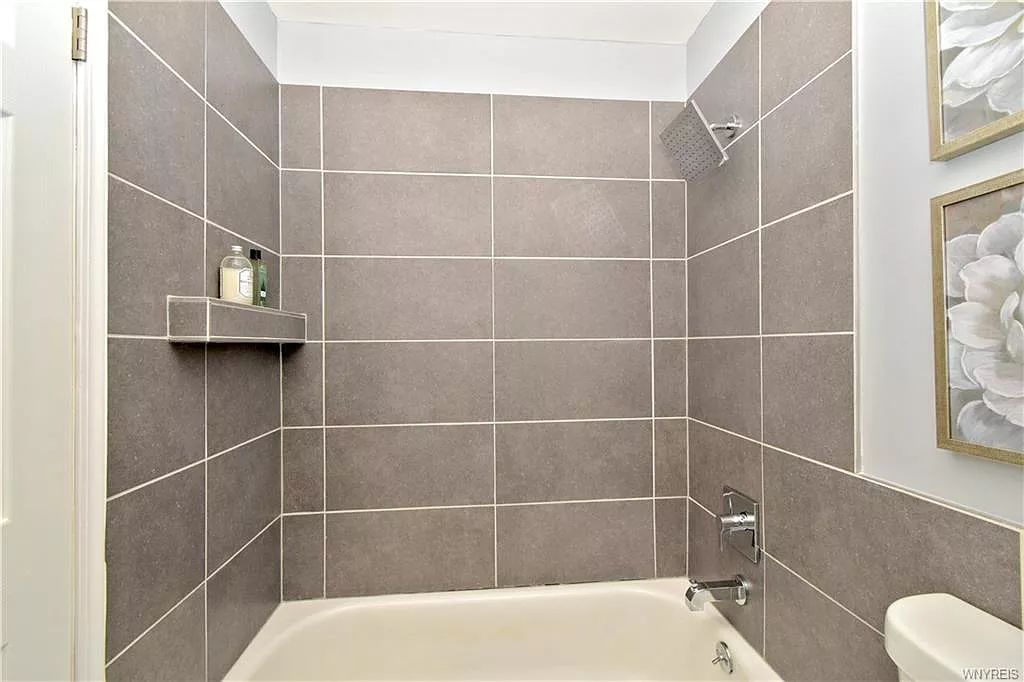 A bathtub and shower combo with neutral gray rectangular tiles, a built-in shower shelf, and two floral wall art pieces.