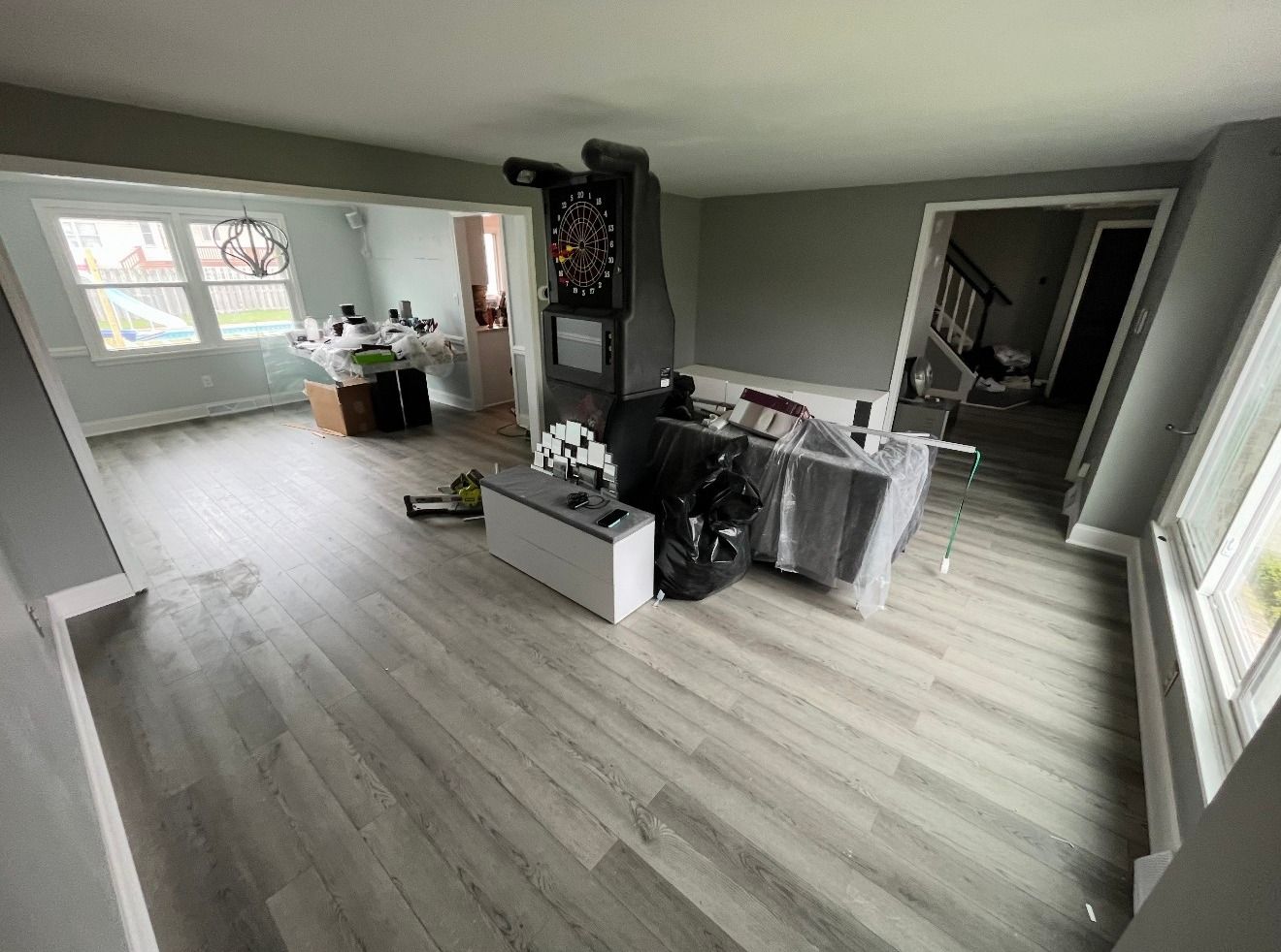 A bright, open-plan living room with light gray walls, wood-look flooring, a central arcade game, and adjacent dining area.