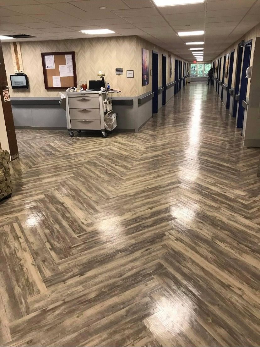A wide, brightly lit hospital hallway with wood-patterned flooring, a nurse's cart, and a bulletin board.