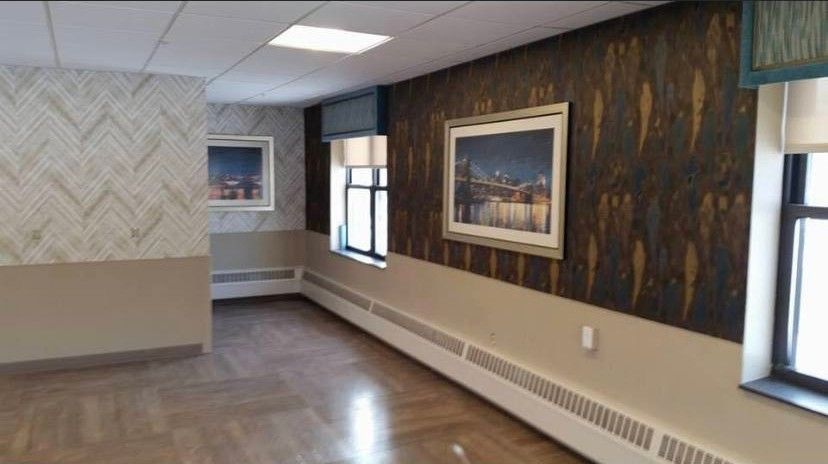 A room with wood floors, two-tone walls with patterned wallpaper, two windows, and two framed wall artworks.