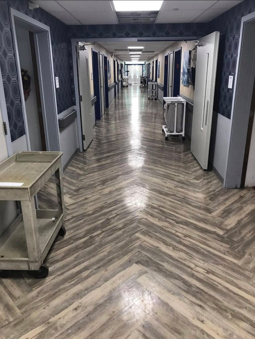 A long, well-lit hospital corridor with herringbone-patterned flooring, blue patterned walls, and medical supply carts.