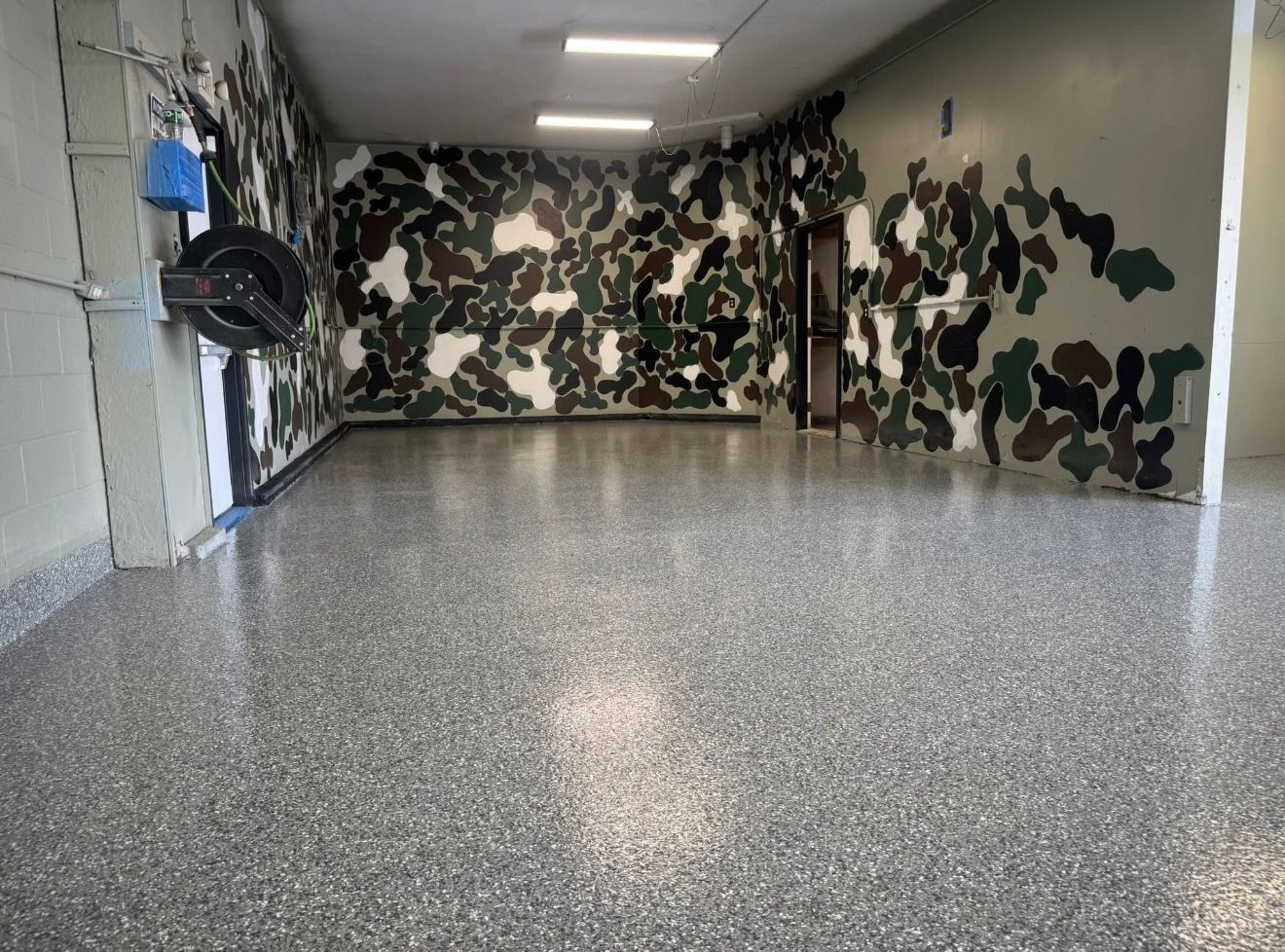 An empty garage with camouflage-painted walls and a glossy grey speckled epoxy floor under bright overhead lights.