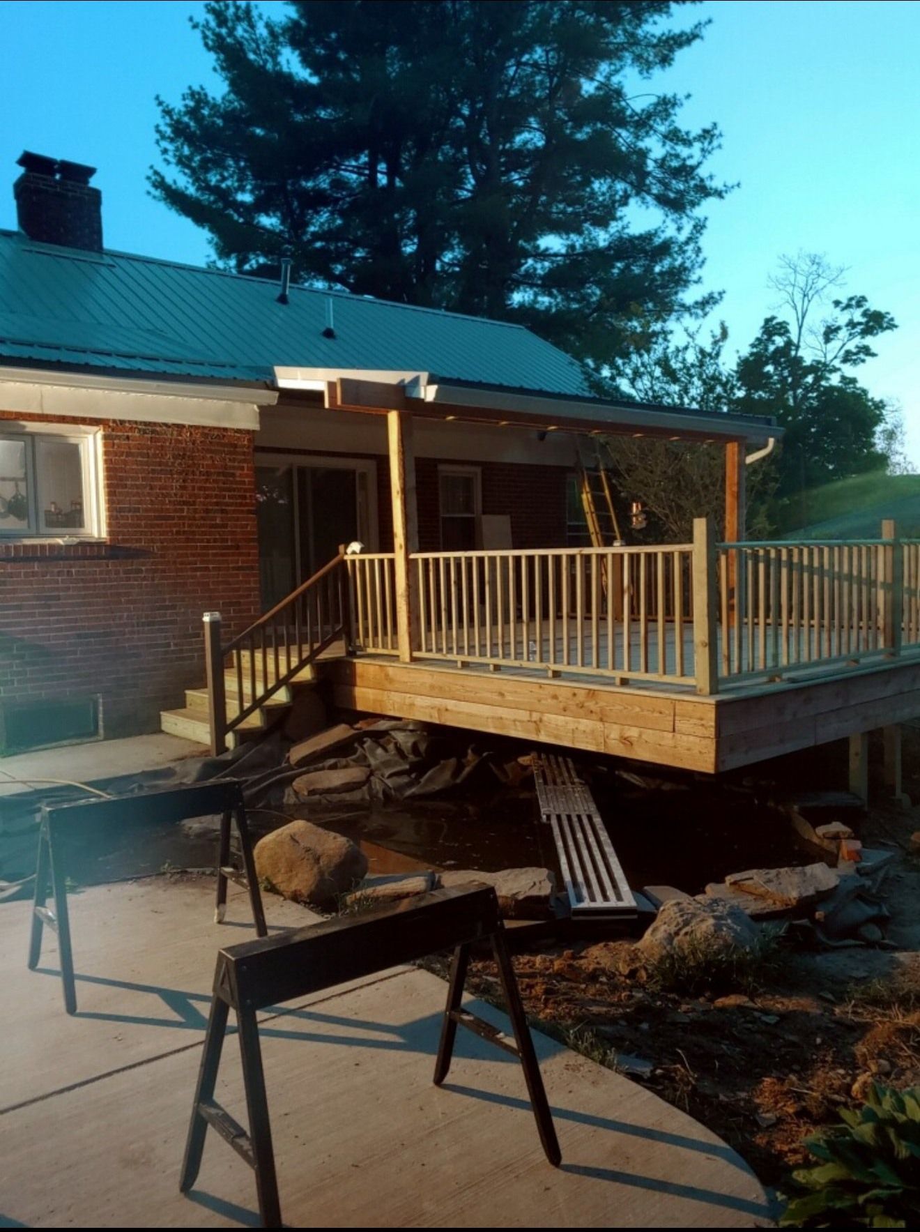 A newly built wooden deck attached to a brick house, overlooking a yard with a concrete patio and two sawhorses.