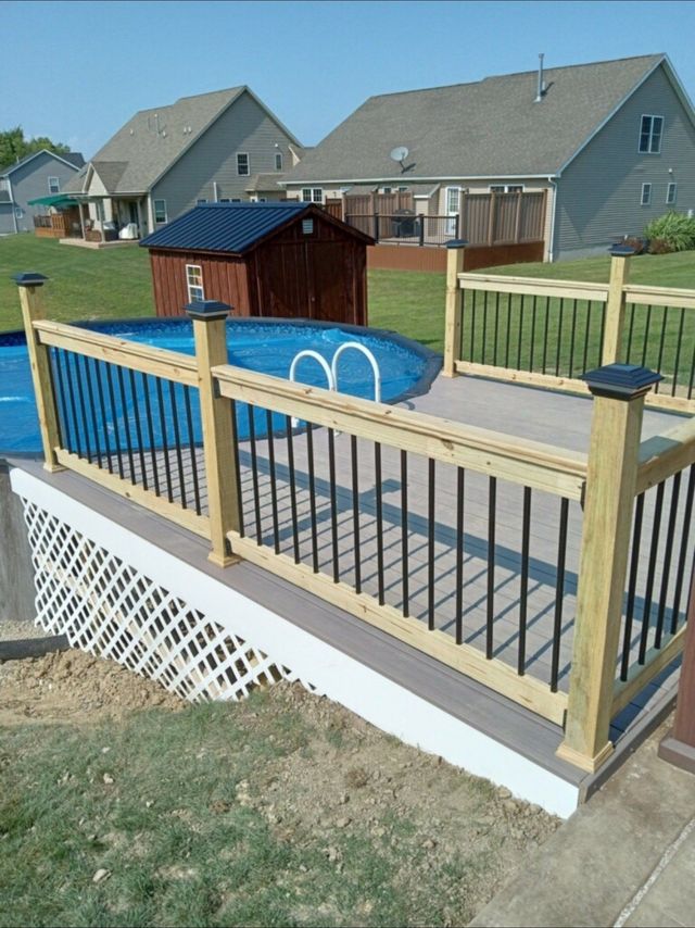 Wooden deck with black railing surrounds an above-ground pool in a backyard on a sunny day.