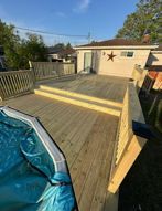 A wooden multi-level deck surrounding an above-ground pool, leading to the entrance of a house with a star wall ornament.