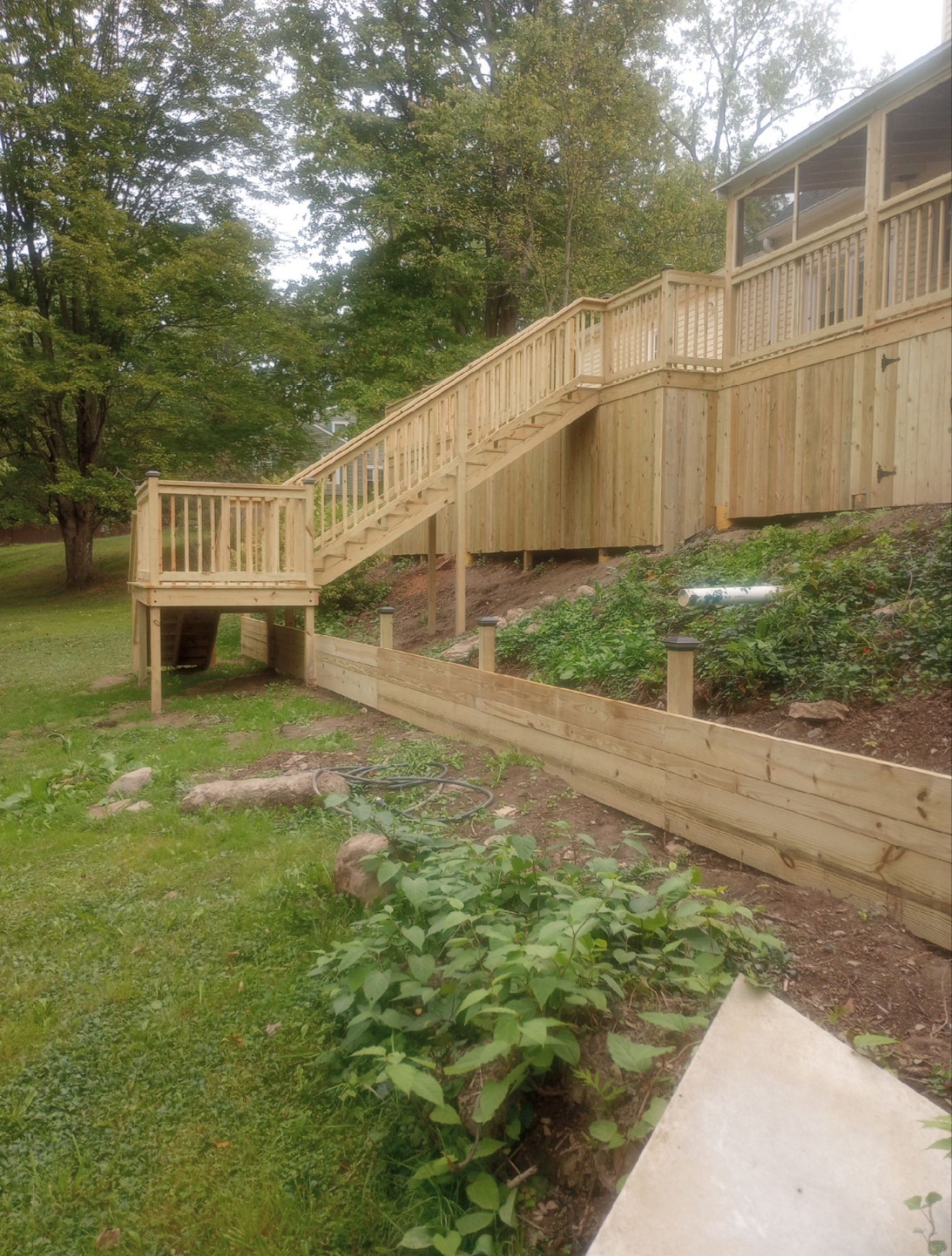 A wooden deck with a staircase leading down to a grass lawn, next to a low wooden retaining wall and garden beds.