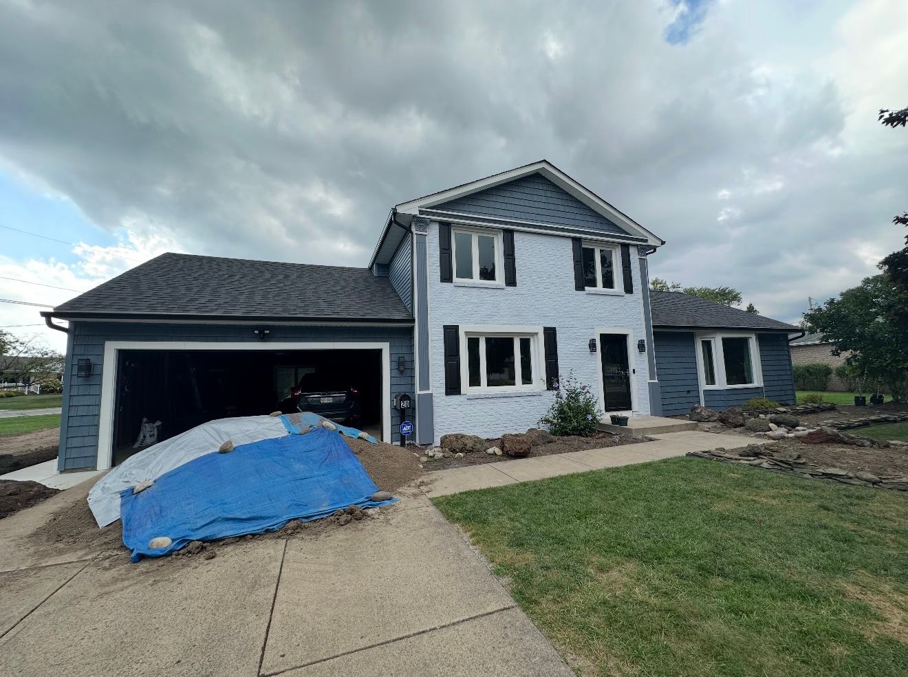 A two-story blue house under a cloudy sky, featuring a garage with a blue tarp in the driveway and some landscaping work.