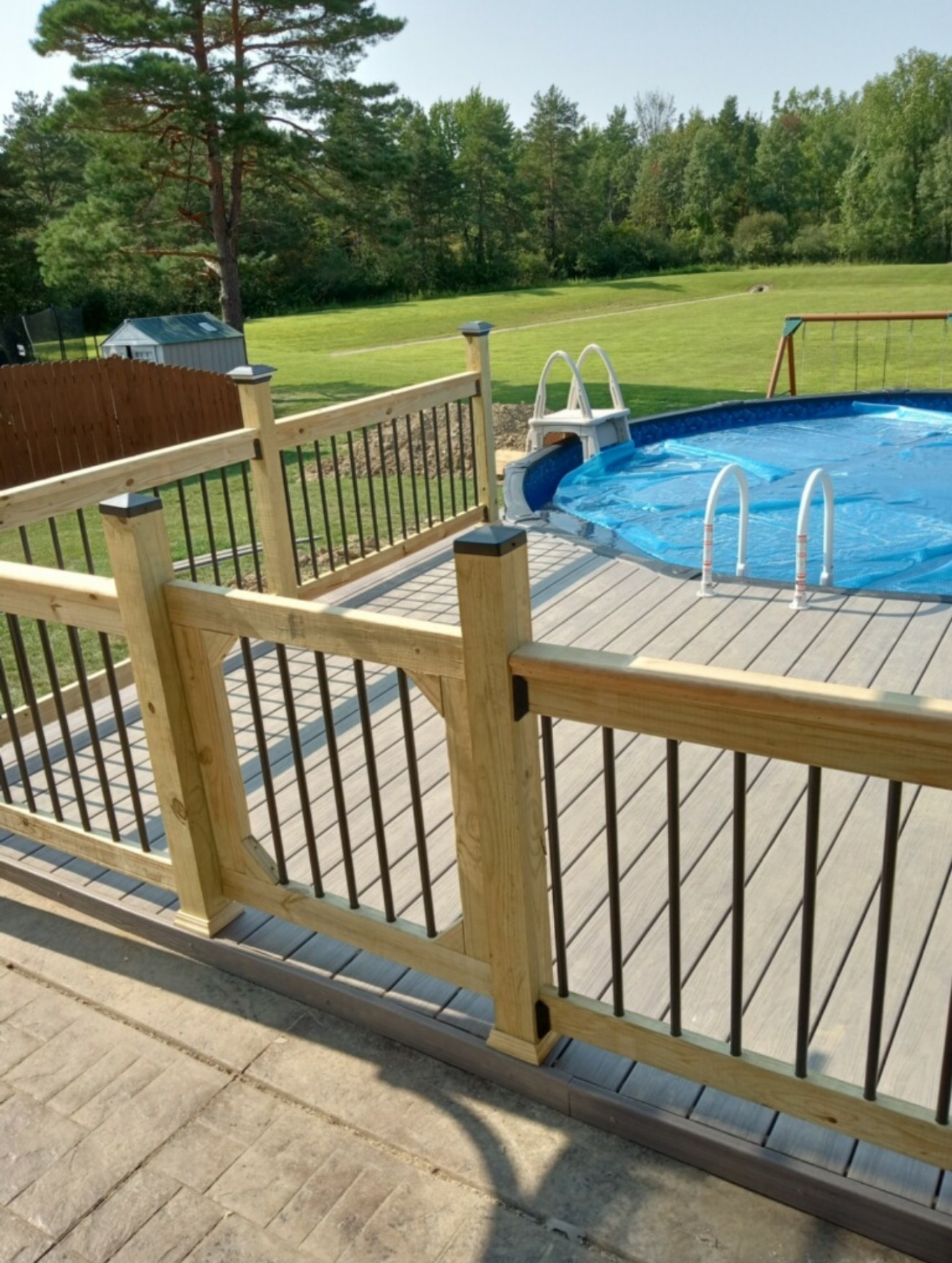 A wood and black metal railing frames a deck with a blue-covered above-ground pool in a grassy backyard.