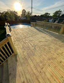 A newly constructed wooden deck featuring railings, overlooking a backyard swimming pool at sunset.