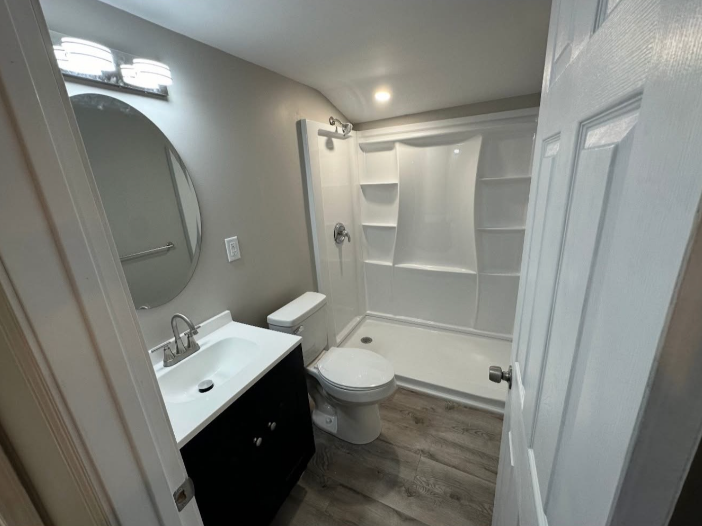 A small, modern bathroom with a black vanity, oval mirror, white walk-in shower, toilet, and grey wood-look flooring.