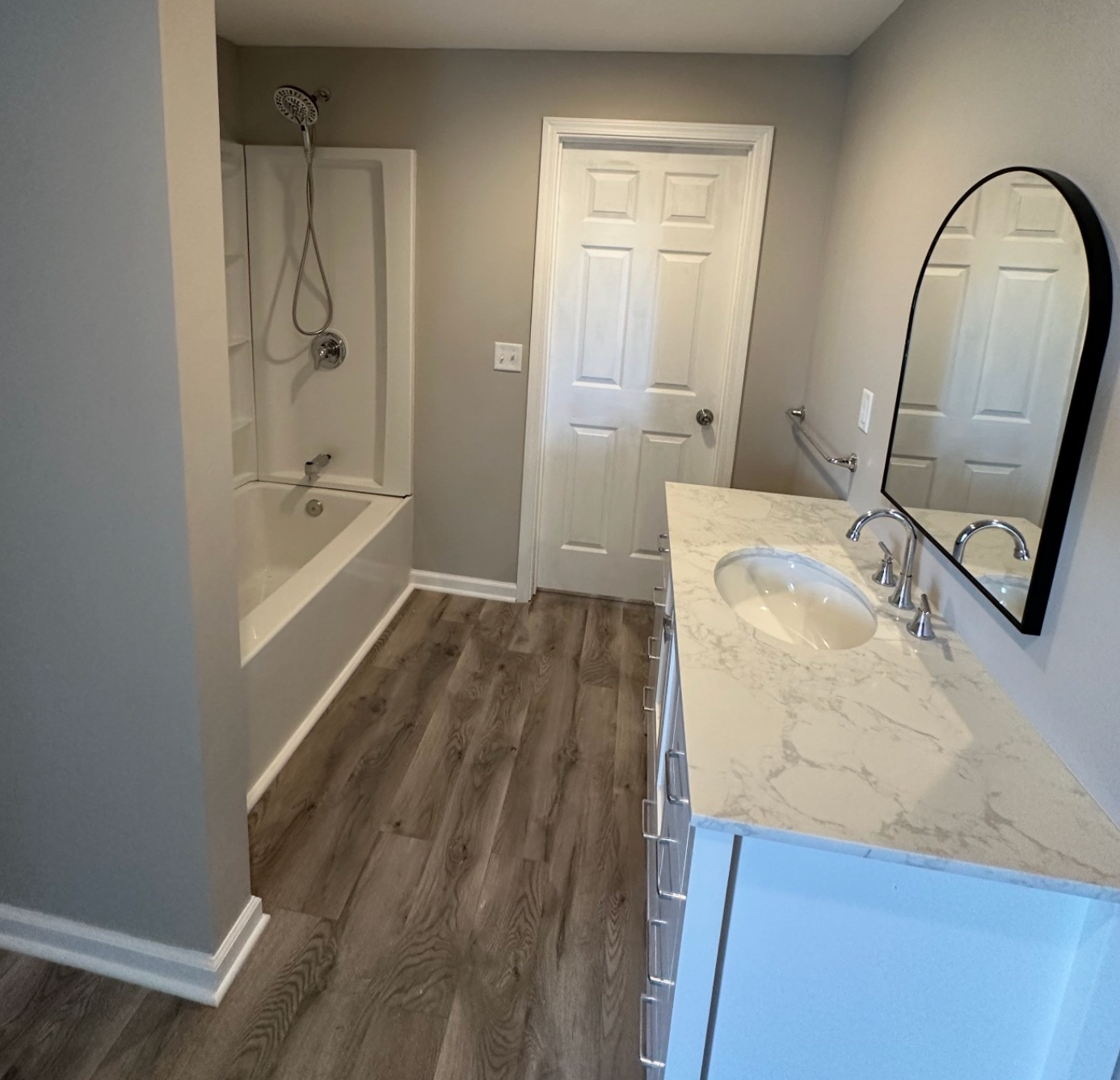 A modern bathroom featuring grey walls, wood-look flooring, a white vanity with a marble top, and a white bathtub shower.