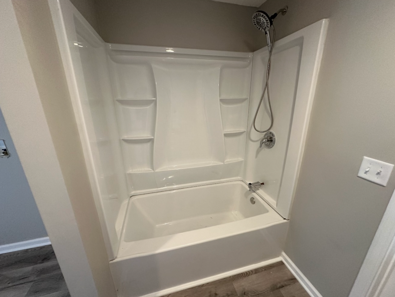 A white fiberglass bathtub and shower surround with a handheld shower head mounted on a grey wall in a bathroom.