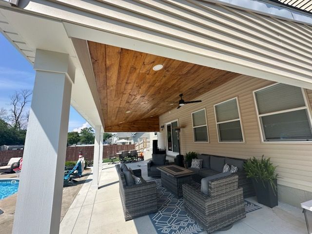 A covered patio with a wooden ceiling, ceiling fan, outdoor wicker seating, and a fire pit, adjacent to a swimming pool.