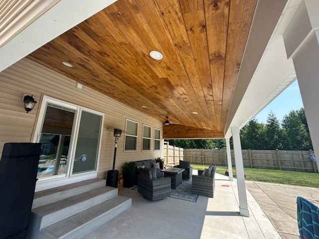 A covered patio with a stained wood ceiling, gray wicker furniture, and concrete stairs leading to a sliding glass door.