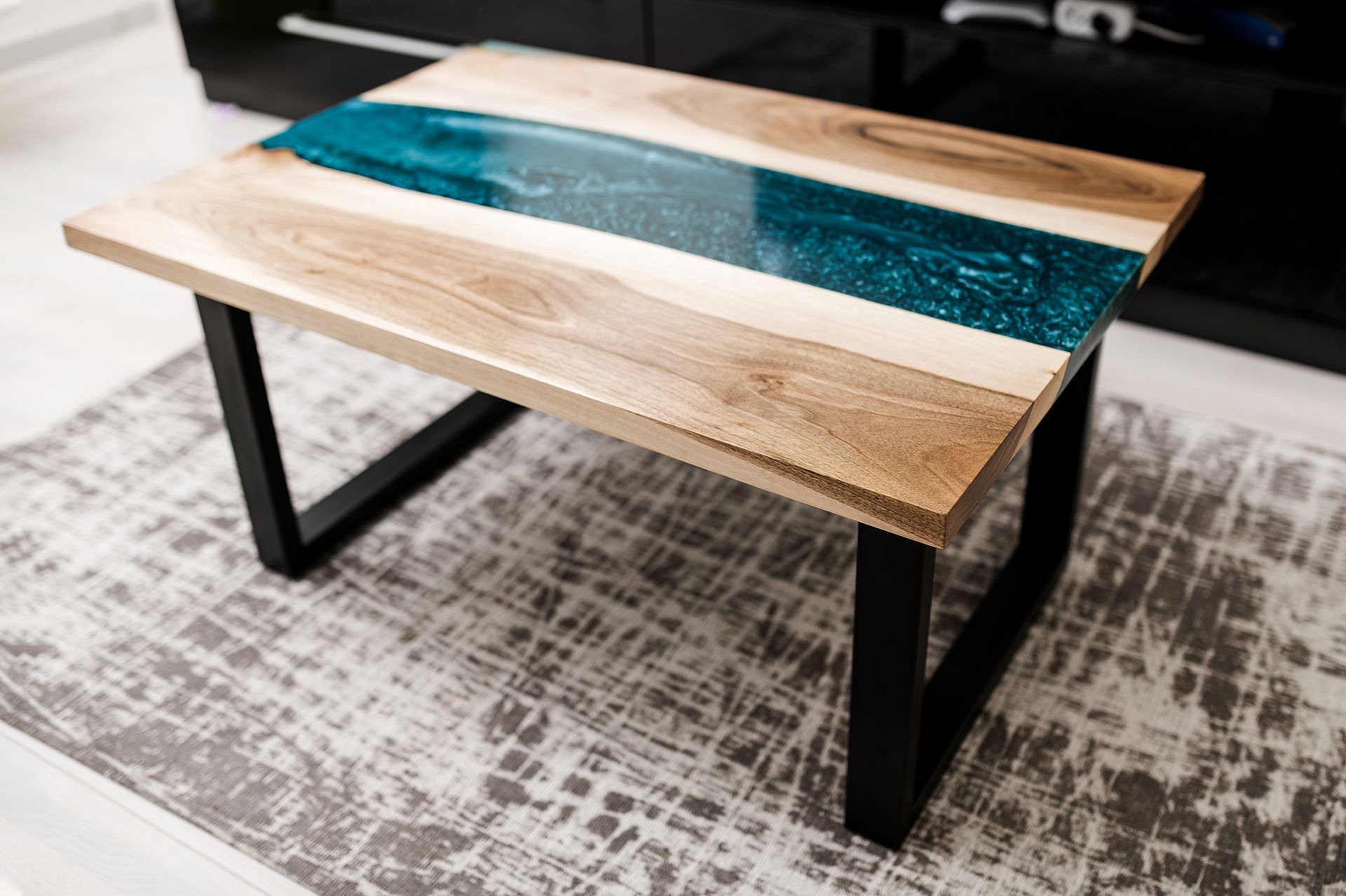 A rectangular coffee table featuring natural wood slabs separated by a glossy, teal resin river, set on black metal legs.