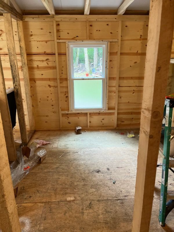 Interior view of a wood-framed room under construction, featuring an installed window and plywood subfloor.