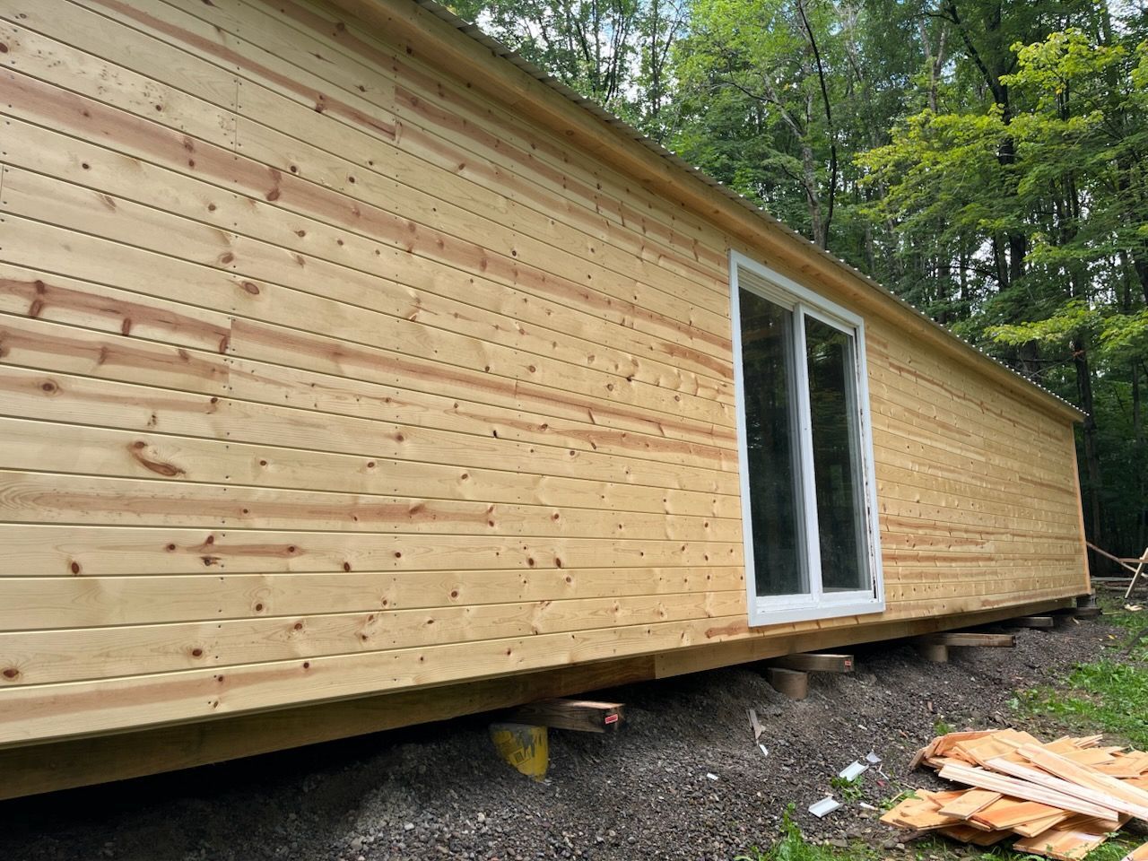 A newly constructed wooden cabin exterior with light wood siding and a white sliding glass door, set on a gravel foundation.