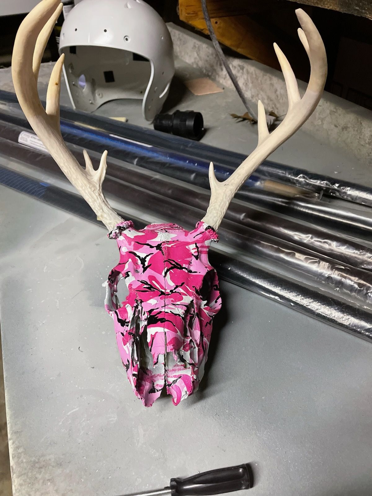 A deer skull with large antlers, painted in a bright pink and black camouflage pattern, sits on a workbench.