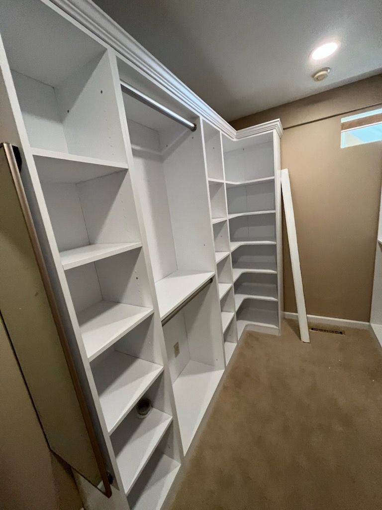 A walk-in closet featuring white modular shelving, vertical storage, and a hanging rod against tan walls and carpet.