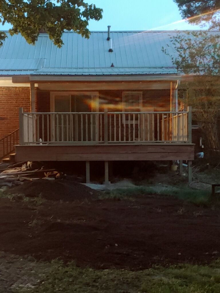 A wooden deck with railings attached to the back of a brick house with a metal roof, overlooking a dirt yard.