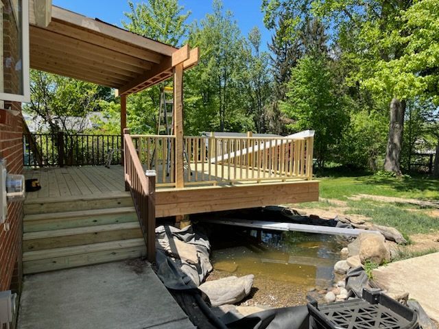 A newly built wooden deck with stairs and a partial roof sits next to a home, overlooking a small, unfinished pond area.