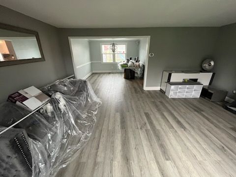 A living room with gray walls, light-wood flooring, a plastic-covered couch, and a white cabinet in the adjacent room.