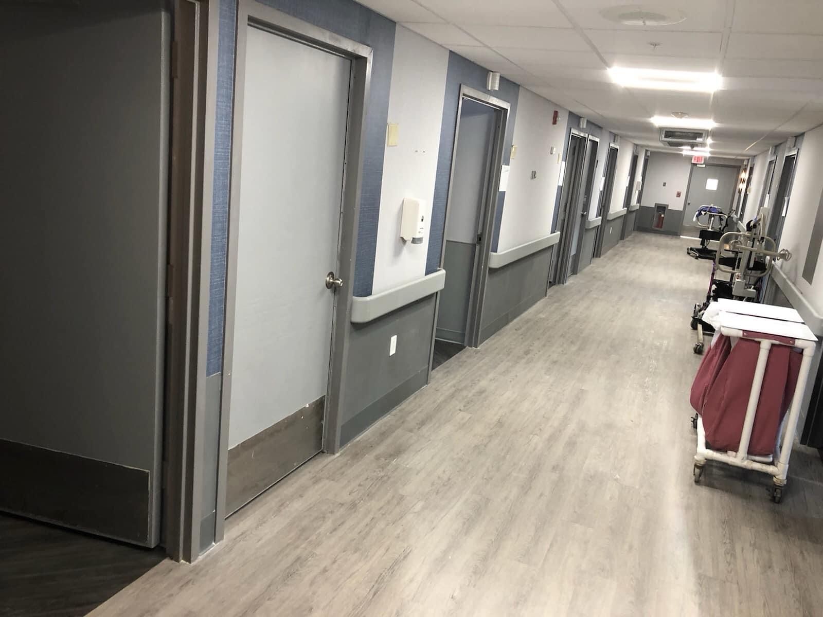 A perspective view of a long hospital hallway with grey doors and a rolling medical cart in the foreground.