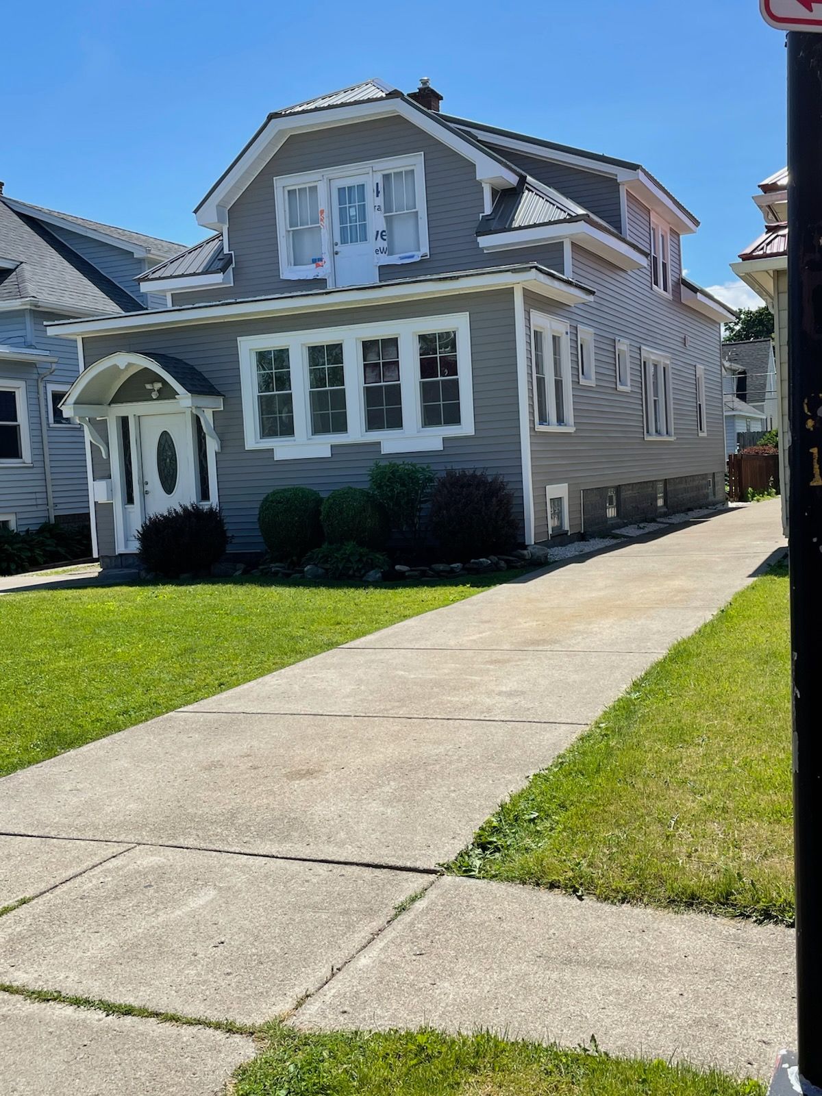 A two-story grey residential house with white trim, a gabled roof, and a concrete driveway on a sunny day.