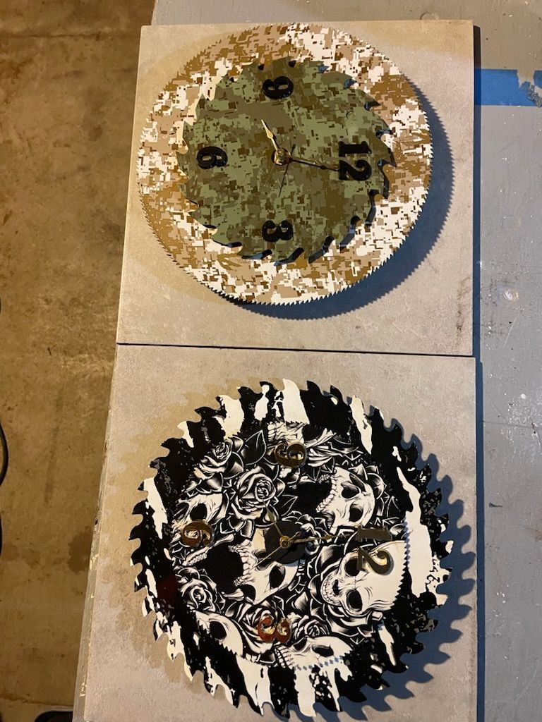 Two circular wall clocks made from saw blades, one with a camouflage pattern and one with a skull design.