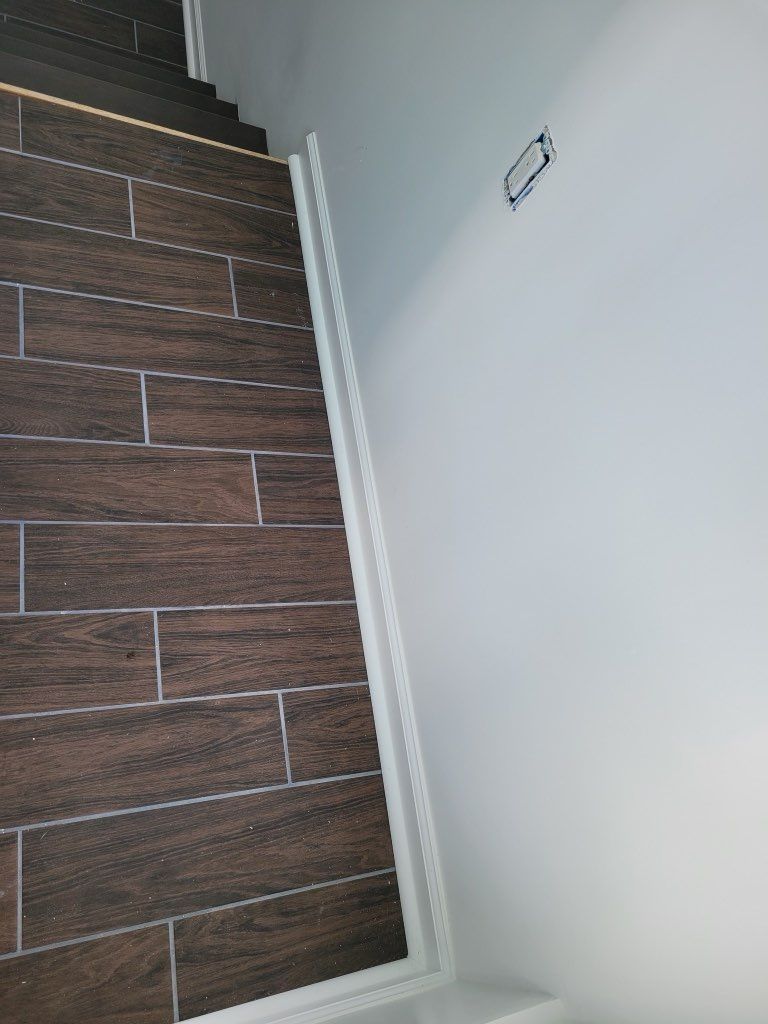 Dark brown wood-look floor tiles meet a white wall with white trim and an electrical outlet box.