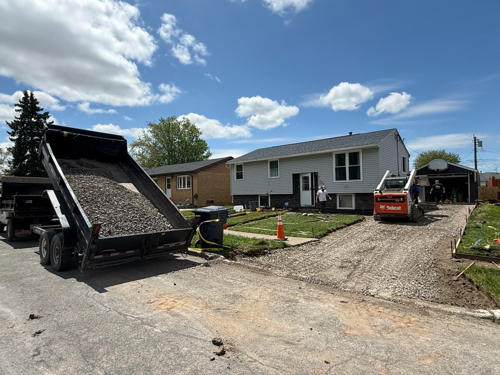 A construction site with a dump truck, a skid steer, and a house with a gravel driveway being prepared for renovation.
