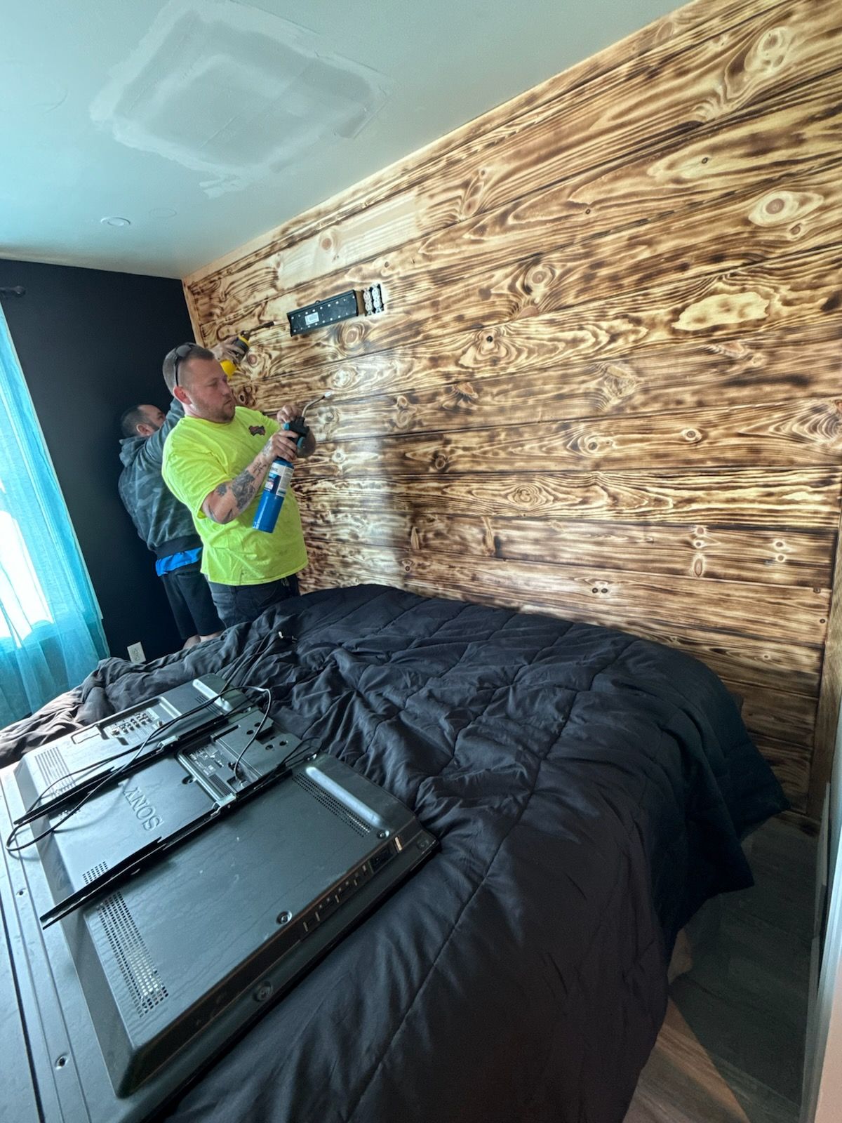 A person in a neon yellow shirt uses a blowtorch on a wood-paneled wall above a bed while another person assists.