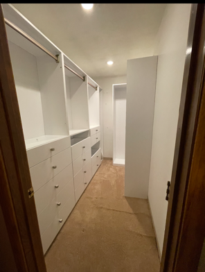 A walk-in closet featuring white modular shelving, hanging rods, and drawers with round knobs on a tan carpet.