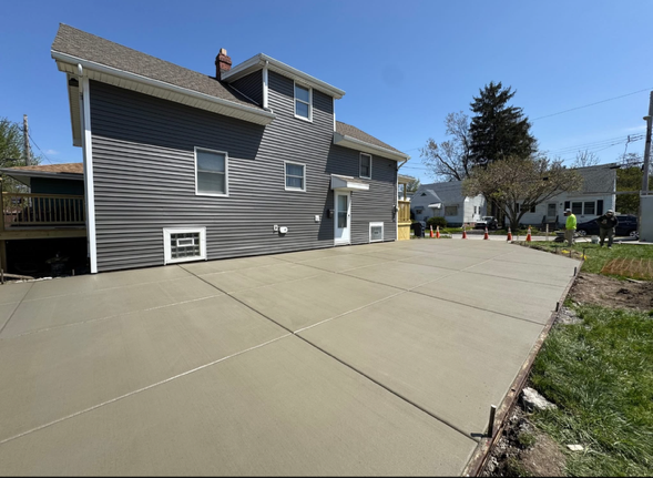 A newly poured concrete driveway extends alongside a two-story gray house under a clear blue sky.