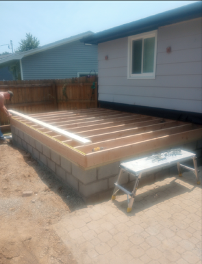 Construction of a wood deck framing on top of a concrete block base next to a gray house.