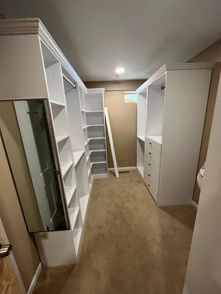 A narrow walk-in closet featuring floor-to-ceiling white shelving units, a hanging rod, drawers, and carpeted flooring.