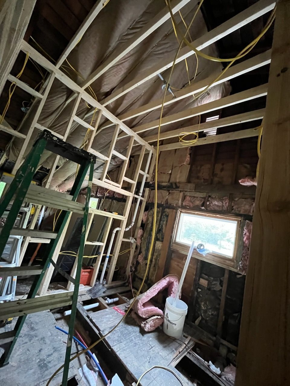 Interior construction scene showing raw wood framing, a window, exposed insulation, electrical wires, and a ladder.